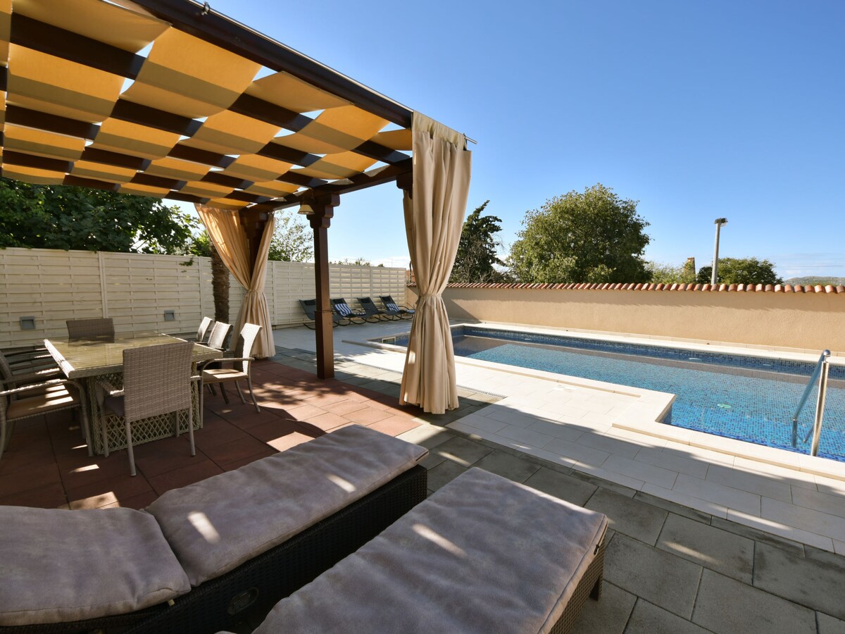 A shaded pergola provides a comfortable dining area with a table and chairs, overlooking a private swimming pool. Lounge chairs are positioned nearby on a spacious patio, surrounded by well-maintained greenery, all under a clear blue sky.