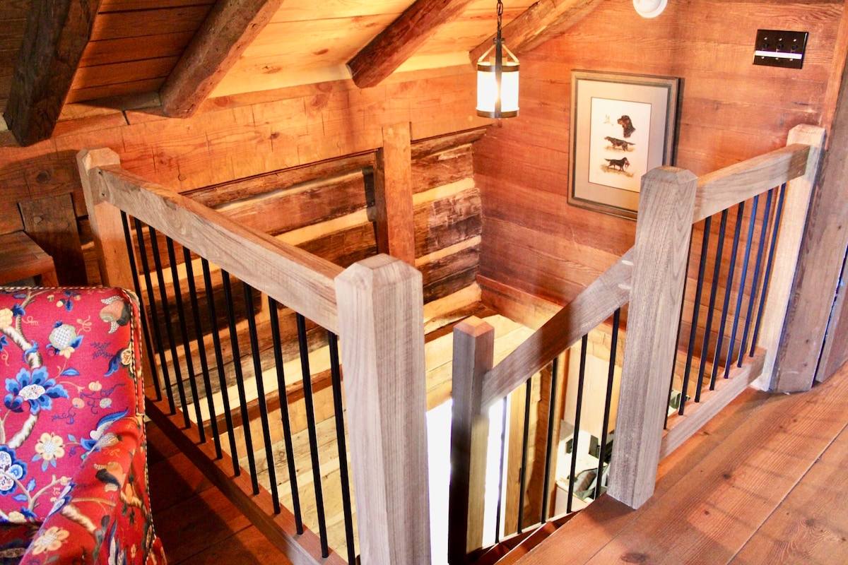 A wooden staircase is framed by rustic posts and metal railings, leading to the second floor. The warm tones of the exposed logs and beams enhance the cabin's charm. A hanging lantern provides soft illumination, while a colorful floral couch is visible on the left.
