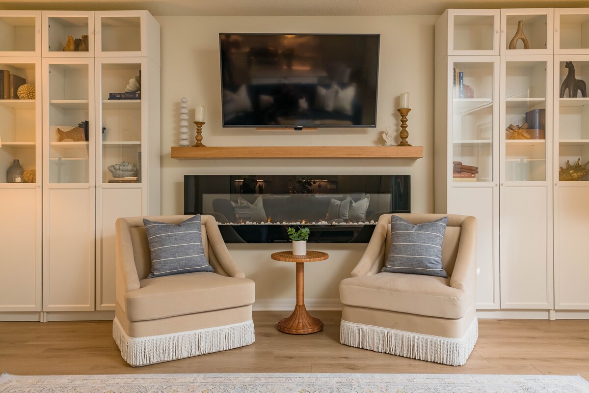 Two cozy chairs with soft blue cushions are positioned facing a modern fireplace, creating a functional relaxation area. A small round table sits between them, adorned with a potted plant. The built-in shelves on either side display decorative items against a neutral wall.