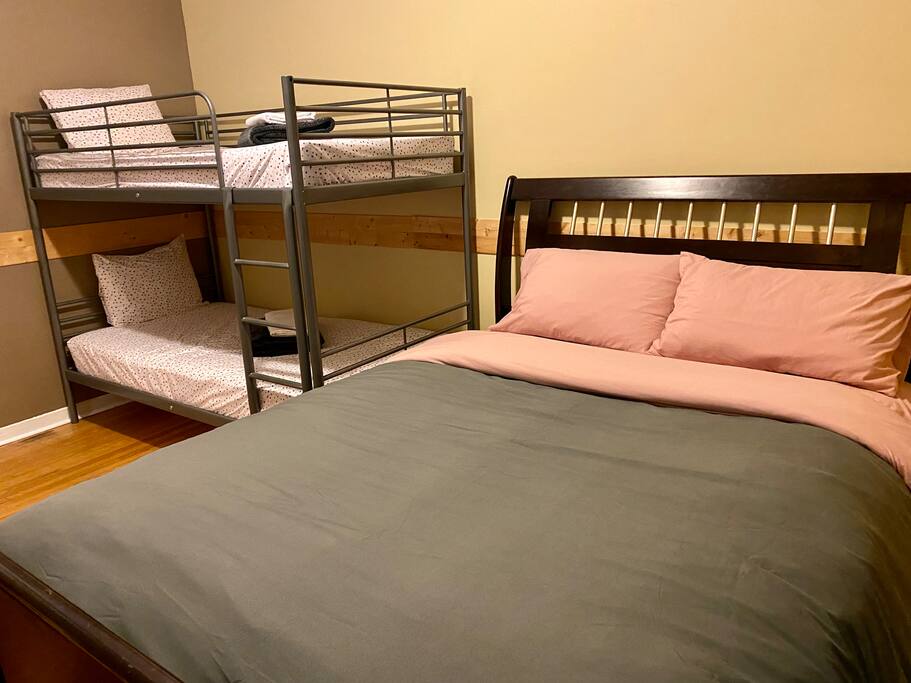 The room features a spacious bed with soft pink bedding and a large frame. Two bunk beds in a metal frame are positioned adjacent, dressed in neutral-toned linens. Light brown walls and hardwood flooring create a welcoming and warm environment.