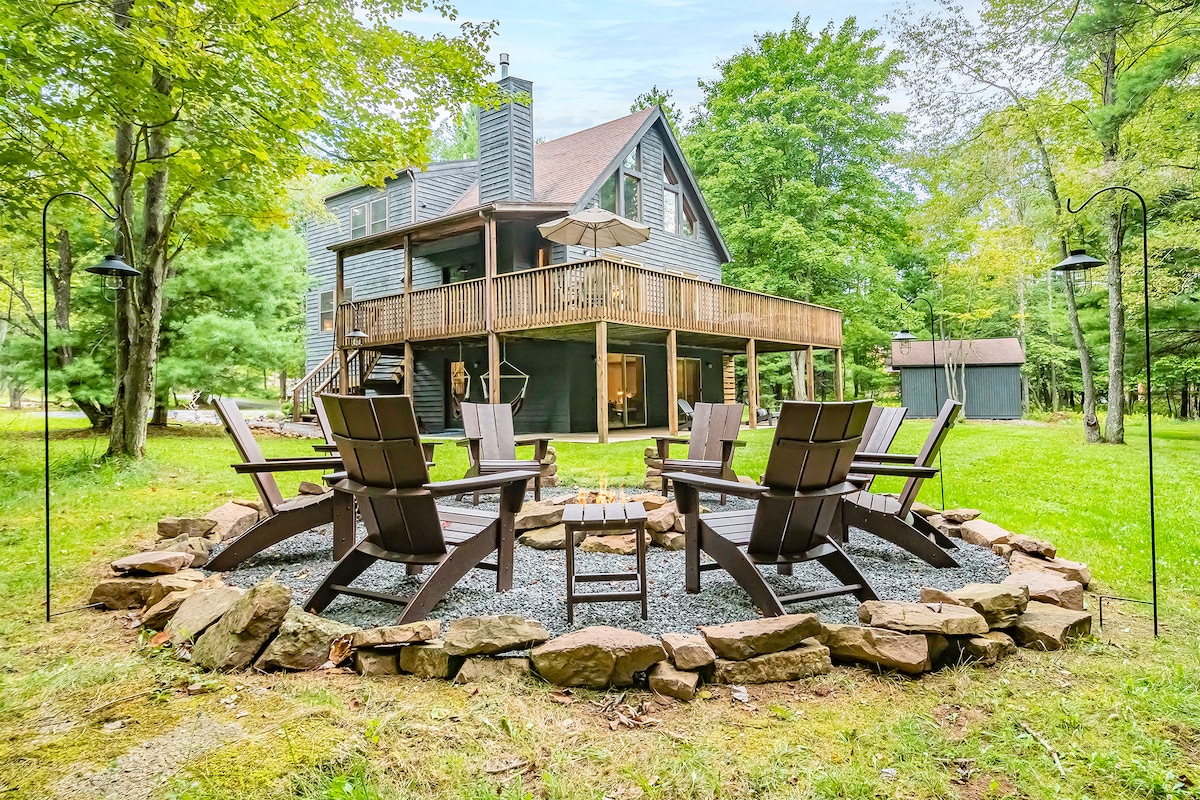 An outdoor fire pit area is surrounded by a circular arrangement of comfortable Adirondack chairs, inviting relaxation. Natural stone borders the pit, complementing the surrounding grassy yard and lush trees. The chalet is visible in the background, showcasing a spacious deck.