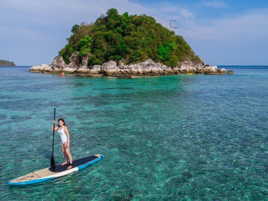 A person stands on a paddleboard, balancing against a backdrop of clear turquoise water. A small, rocky island covered with lush greenery is visible in the distance, with gentle waves lapping at the shore. The serene scene showcases the natural beauty of the surrounding area.