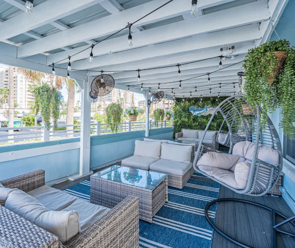 An outdoor lounge area is furnished with plush seating on a stylish rug, inviting relaxation. A glass-top table is centered within the space, while hanging plants add greenery. Soft lighting fixtures are suspended from the ceiling, enhancing the atmosphere of the inviting setting.