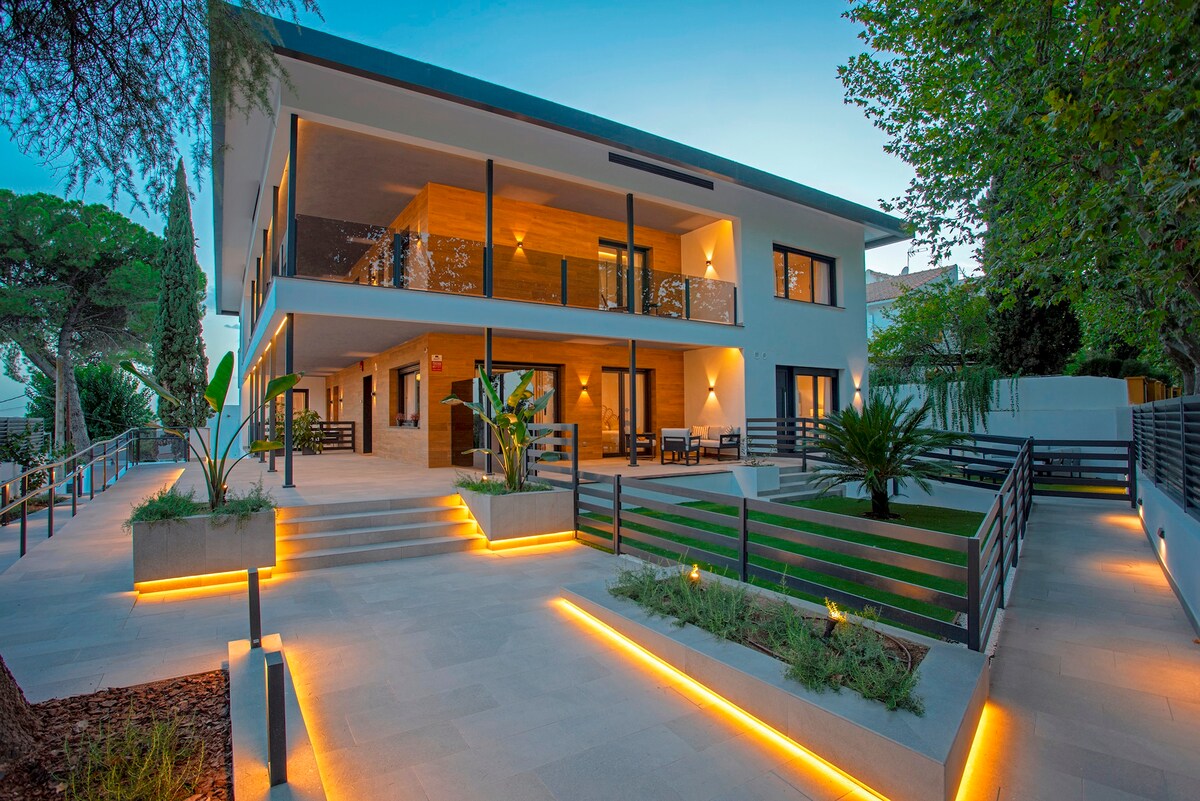 The exterior of a modern building is shown, featuring multiple levels with spacious balconies and large glass doors. A well-maintained garden surrounds the property, enhanced by strategically placed lighting along the pathways. Seating areas are visible on the upper terrace, inviting relaxation.