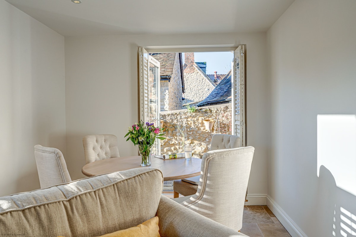 A light-filled dining area features a round table with seating for four. Large windows are opened, allowing fresh air and a view of the outdoor surroundings. A bouquet of flowers adds a touch of color to the inviting space.