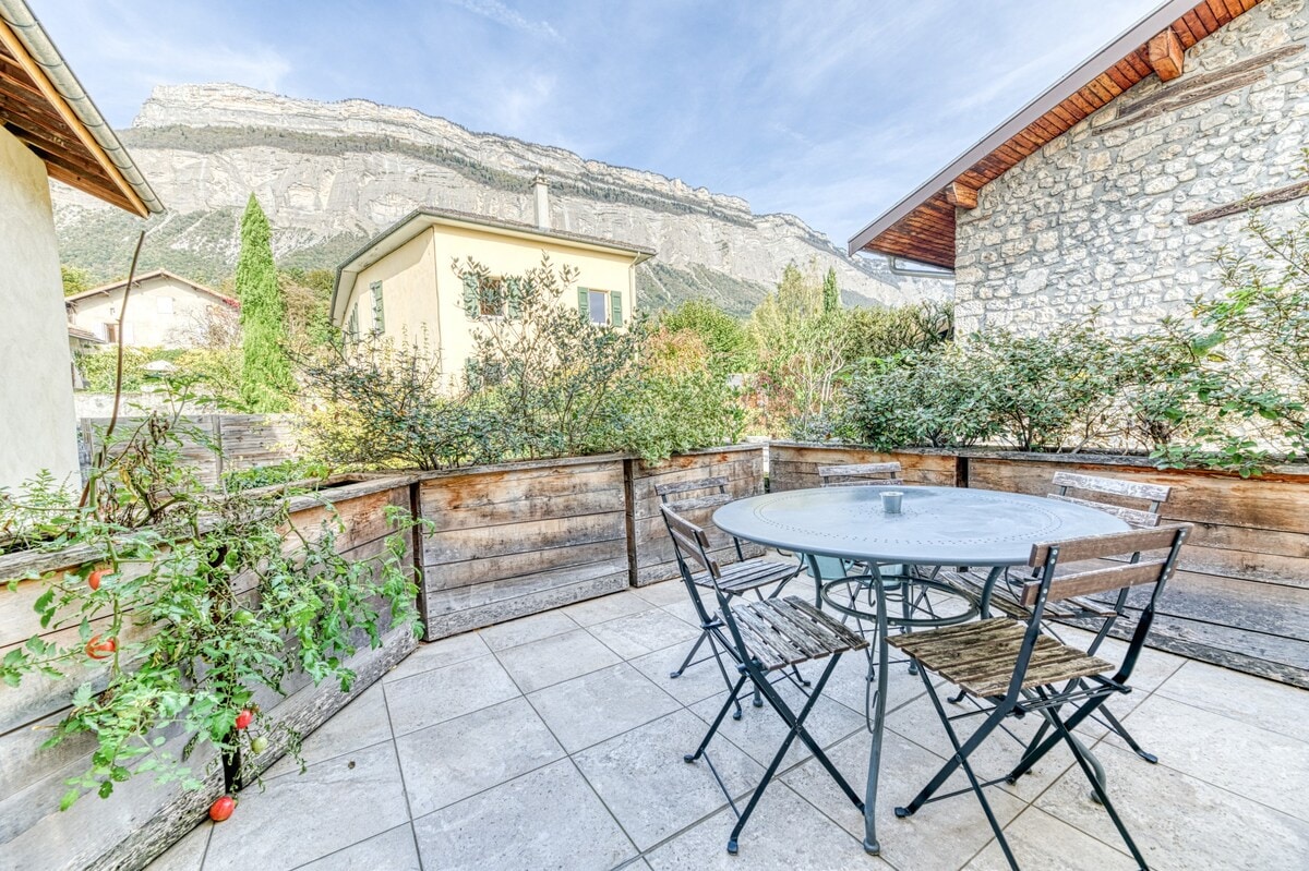 An outdoor terrace features a circular table surrounded by several metal chairs. Lush greenery and low wooden barriers create a natural border, while the majestic mountains rise in the background, providing a scenic view. The area is paved with stone tiles.