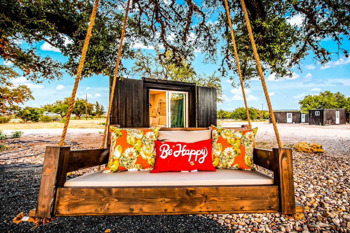 A wooden swing bed is surrounded by vibrant floral cushions and a decorative pillow that reads 'Be Happy.' In the background, shipping containers are visible among the expansive outdoor space, framed by lush trees and a clear blue sky.