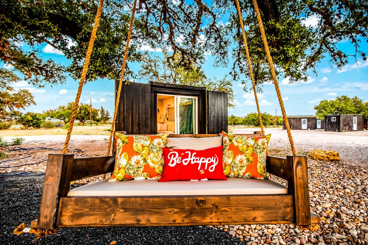 A wooden swing adorned with colorful, floral pillows is suspended from a large tree. A bright 'Be Happy' pillow adds a cheerful touch. In the background, the entrance to a shipping container home is visible, surrounded by gravel and greenery.