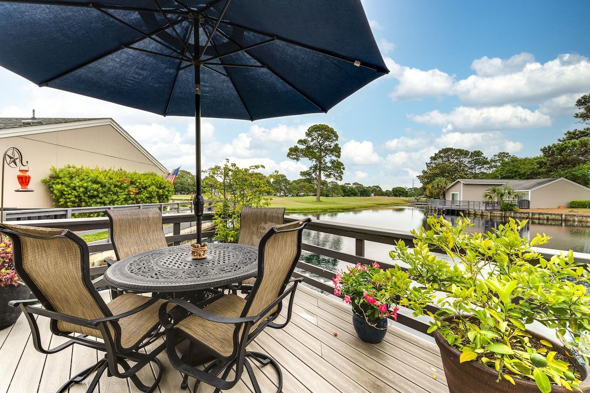 An outdoor deck is presented with a round table and six chairs under a blue umbrella. Potted plants add greenery, while a calm retention pond is visible in the background, with trees and light clouds enhancing the serene environment.