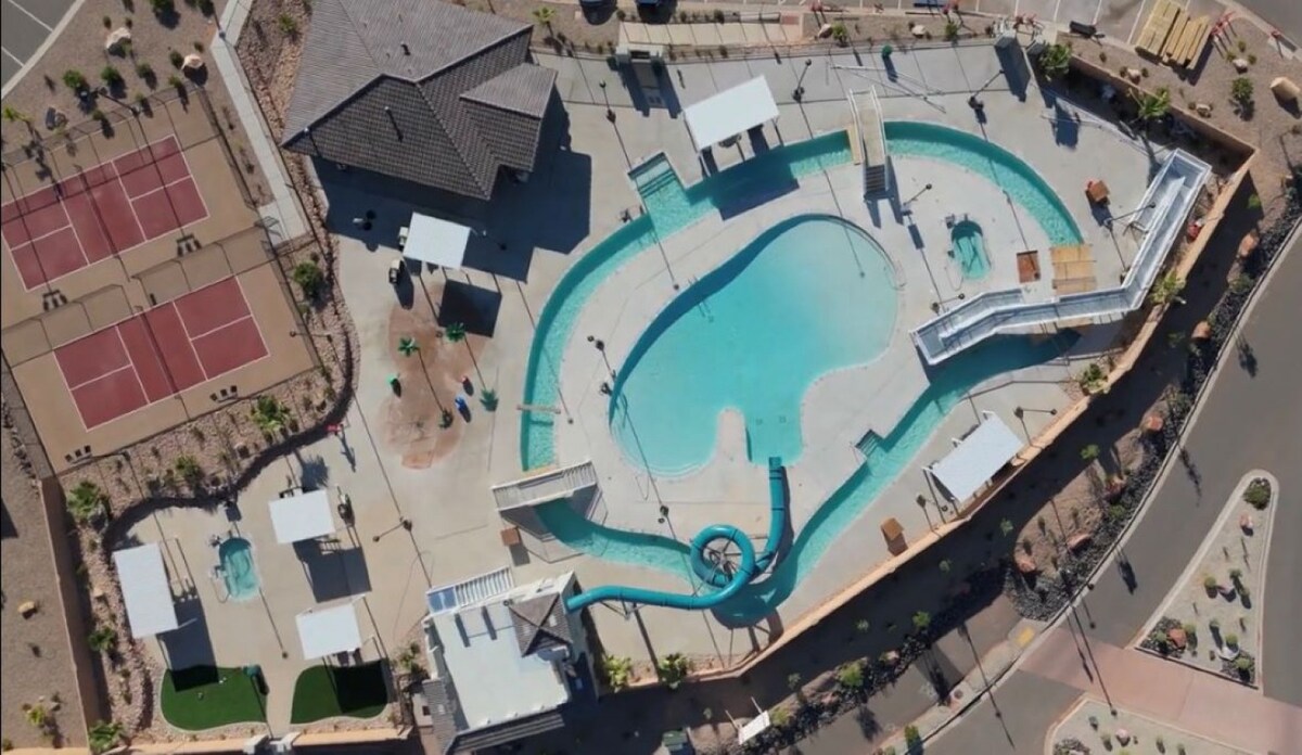 An aerial view of the community amenities reveals a large pool area with a water slide, surrounded by lounging areas. Two tennis courts are visible nearby, along with shaded seating areas, showcasing a well-designed outdoor space for relaxation and recreation.