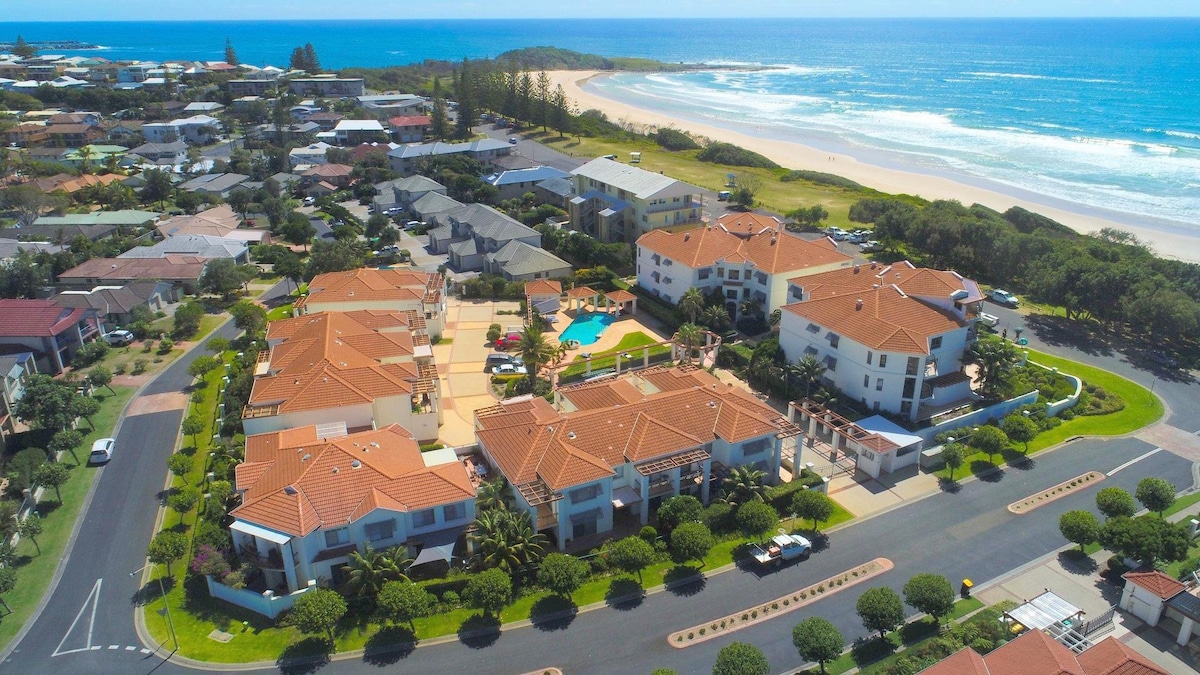 An aerial view captures the resort layout with several buildings and a central pool area highlighted. The coastline runs parallel, showcasing sandy beaches and lush greenery surrounding the property. The neighborhood features residential homes and well-maintained roads leading to the beach.