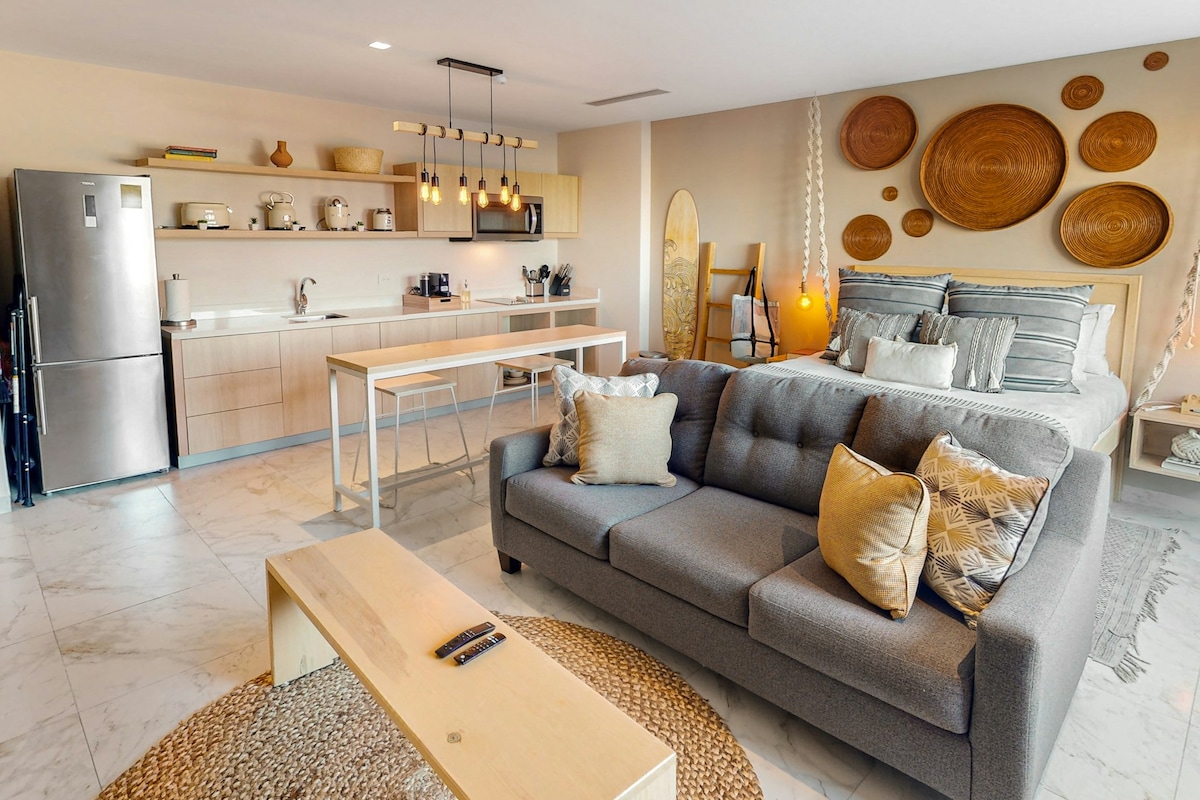 The interior of the studio displays an open layout, featuring a modern kitchen and cozy living area. A spacious bed is positioned near decorative wall art, while a light-colored coffee table sits on a woven rug in front of a plush sofa adorned with cushions.