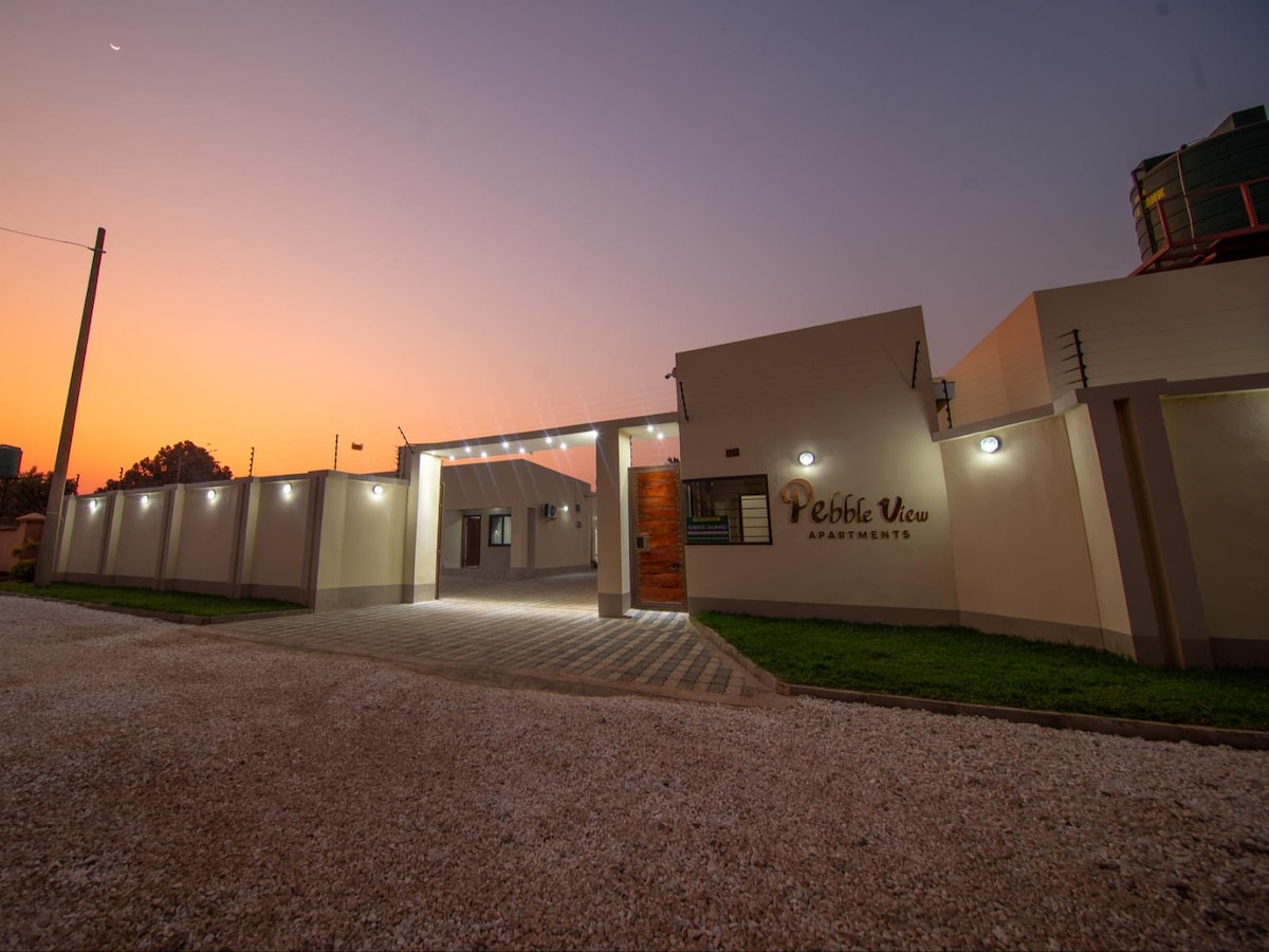The exterior of the apartment complex is displayed at dusk, featuring a welcoming entrance illuminated by soft lighting. A gravel driveway leads up to the entrance, surrounded by neatly trimmed grass. The building's name, 'Pebble Vex Apartments,' is prominently featured above the door.