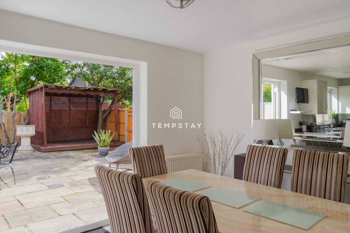 A dining area features a spacious wooden table surrounded by striped chairs. Natural light pours in through large glass doors, connecting the indoor space to an outdoor patio with a gazebo and greenery. A full-length mirror reflects the inviting setting.