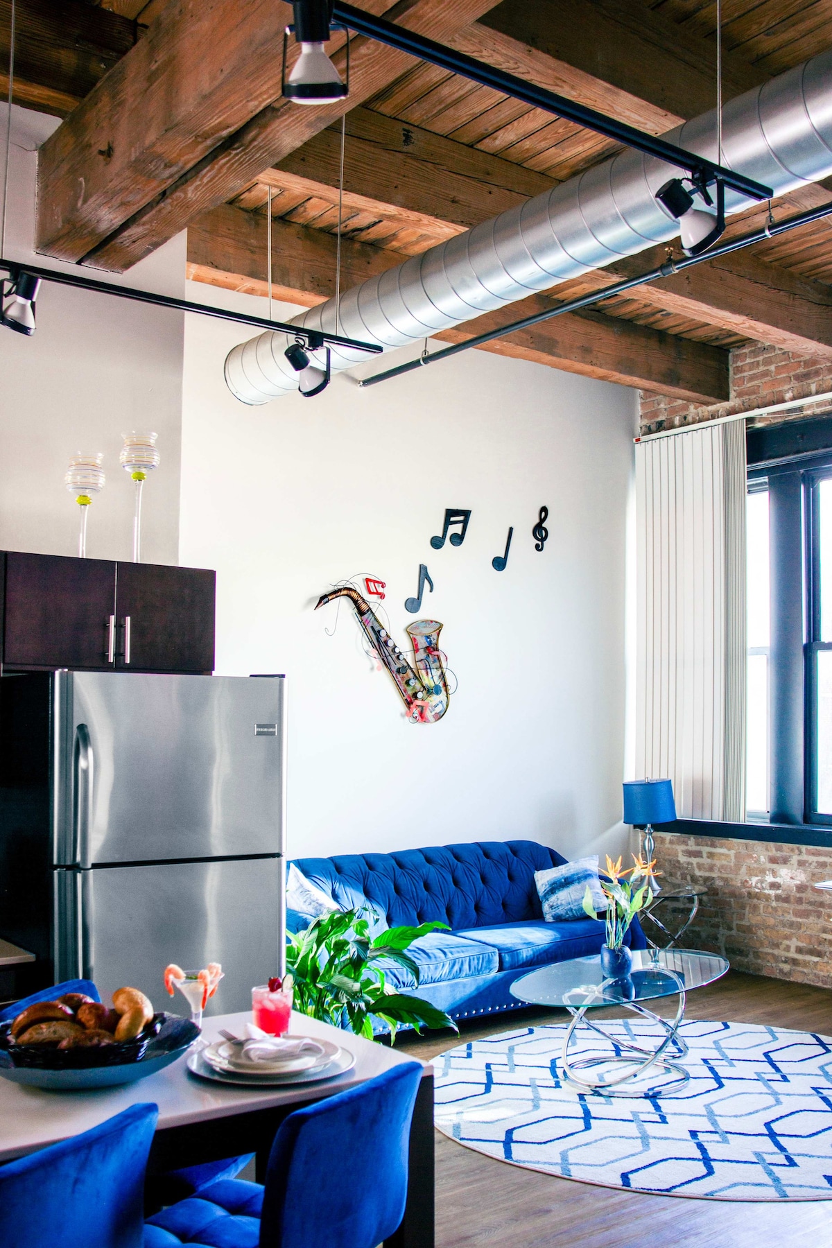 A modern living space features a blue sofa set against a brick wall, complemented by a glass coffee table. The kitchen area includes a silver refrigerator and a dining table adorned with breakfast items. Decorative music notes hang above, enhancing the lively ambiance.