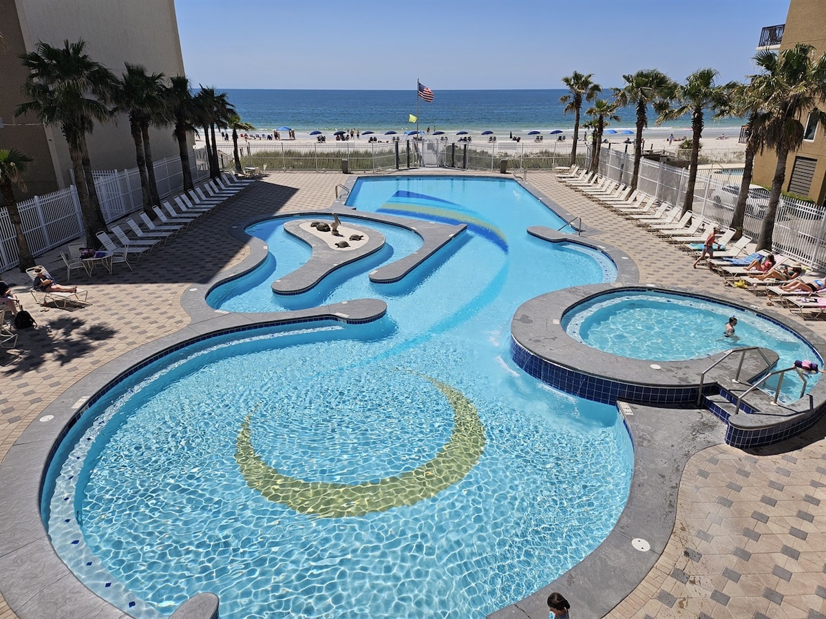 An expansive beachside pool area features a winding layout with integrated hot tubs. Palm trees line the perimeter, while views of the Gulf of Mexico are visible in the background. Lounge chairs are arranged along the deck, inviting relaxation by the water.