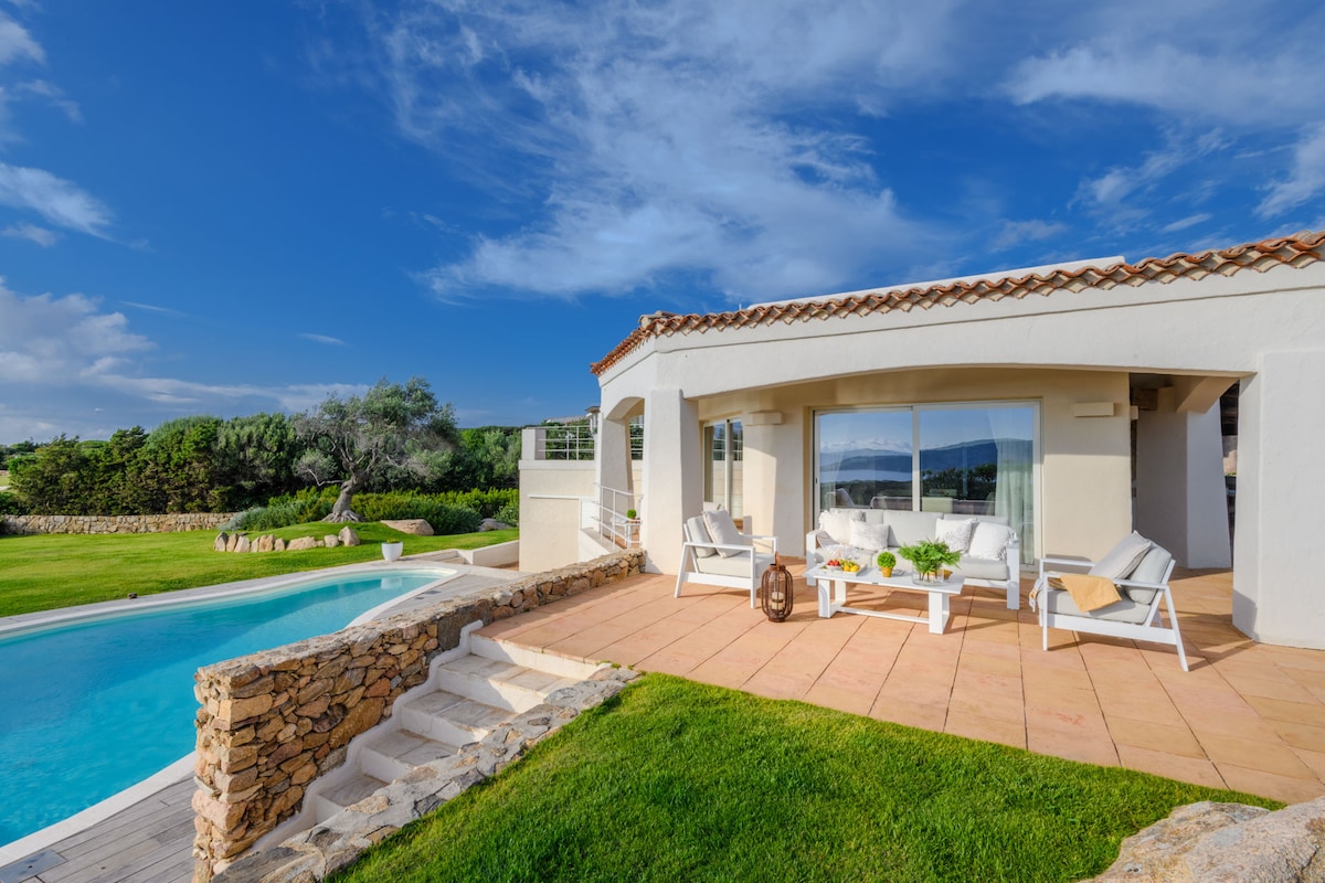 A spacious outdoor terrace features white lounge chairs accompanied by a small table and a vase of fresh fruit. Stone steps lead to a shimmering swimming pool surrounded by lush green grass and gardens, under a partly cloudy blue sky.