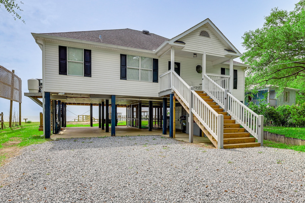 A raised seaside cottage is presented, featuring a welcoming front staircase leading to the main entrance. The exterior showcases a light-colored façade with evenly spaced windows, and a gravel driveway accommodates parking beneath the home. Surrounding greenery adds a natural touch to the setting.