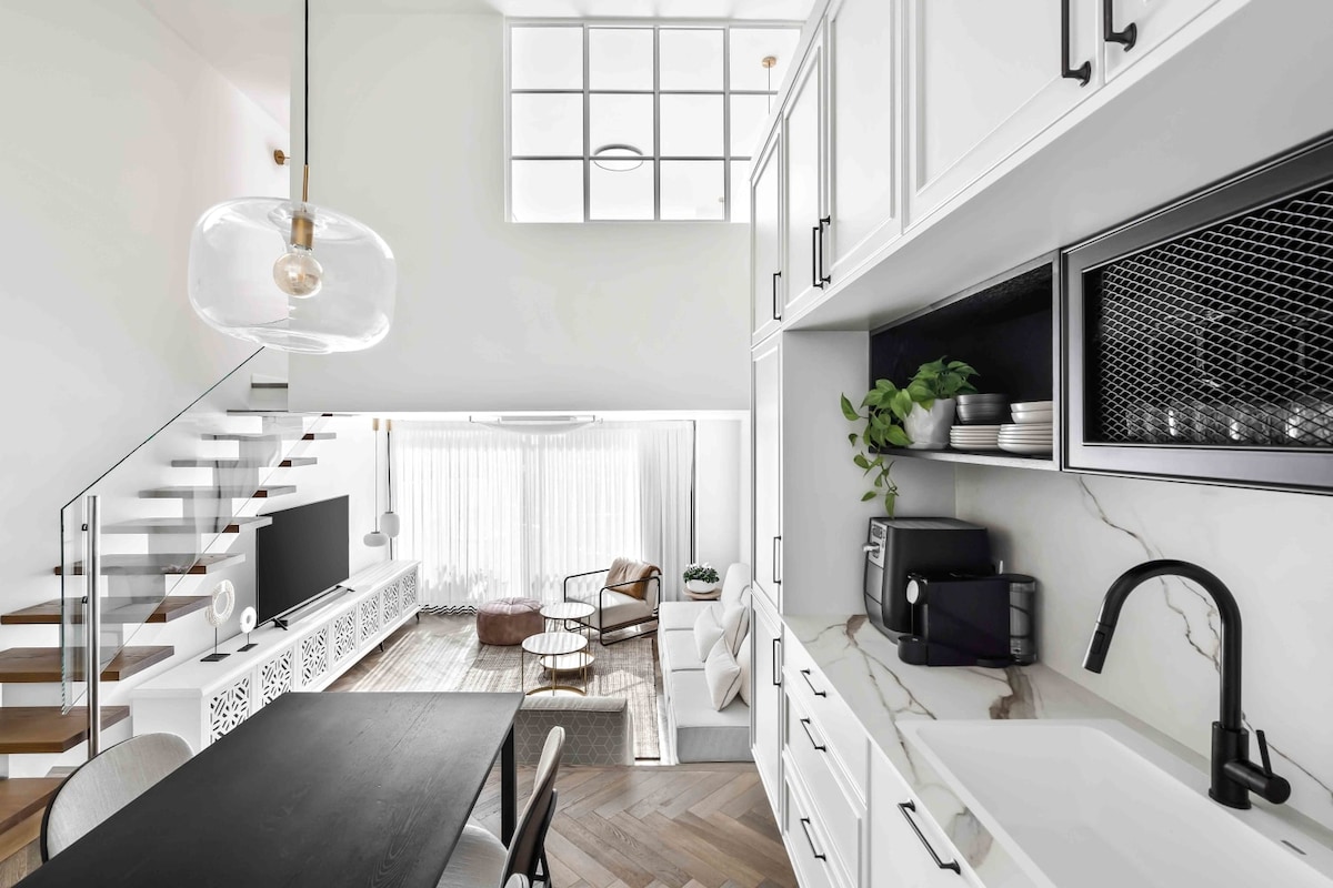A modern kitchen features sleek white cabinetry with open shelving displaying potted plants. The dining area includes a dark table and is illuminated by a pendant light. In the background, a spacious living room is visible, showcasing a light sofa and a large TV surrounded by natural light filtered through sheer curtains.