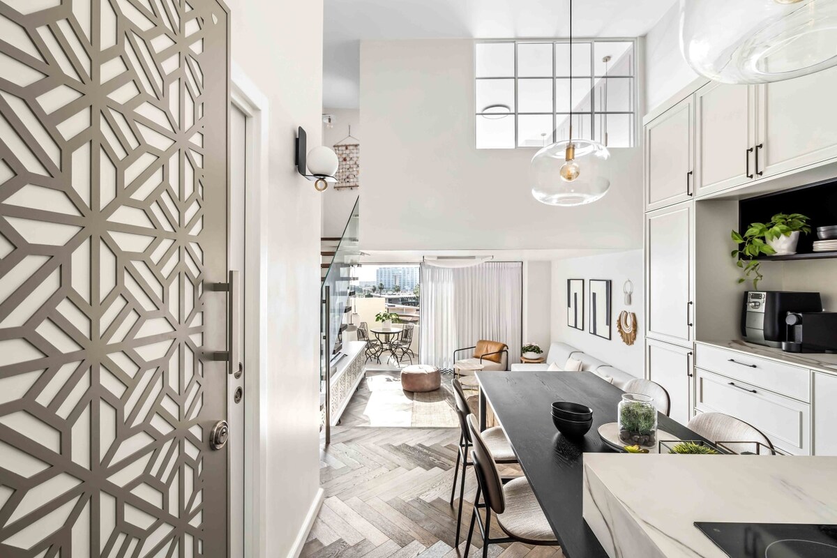 An open kitchen and dining area is presented, featuring a modern design with a black dining table surrounded by light-colored chairs. A geometric-patterned door offers an elegant entryway, while large windows allow daylight to fill the space, accentuating the sleek cabinetry and contemporary décor.
