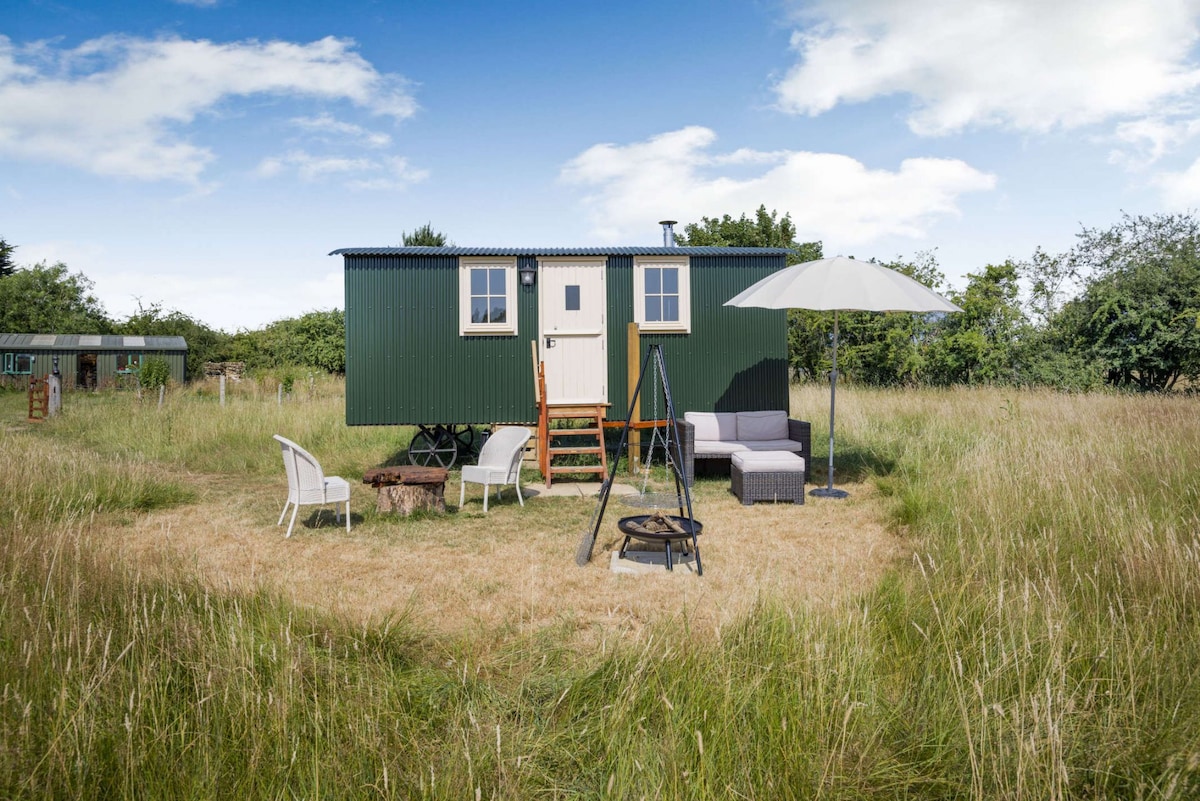 The shepherd's hut is surrounded by tall grasses, with wooden steps leading up to its entrance. A sitting area with small rattan furniture and a parasol is located nearby, along with a firepit in the grassy space, ideal for outdoor relaxation.