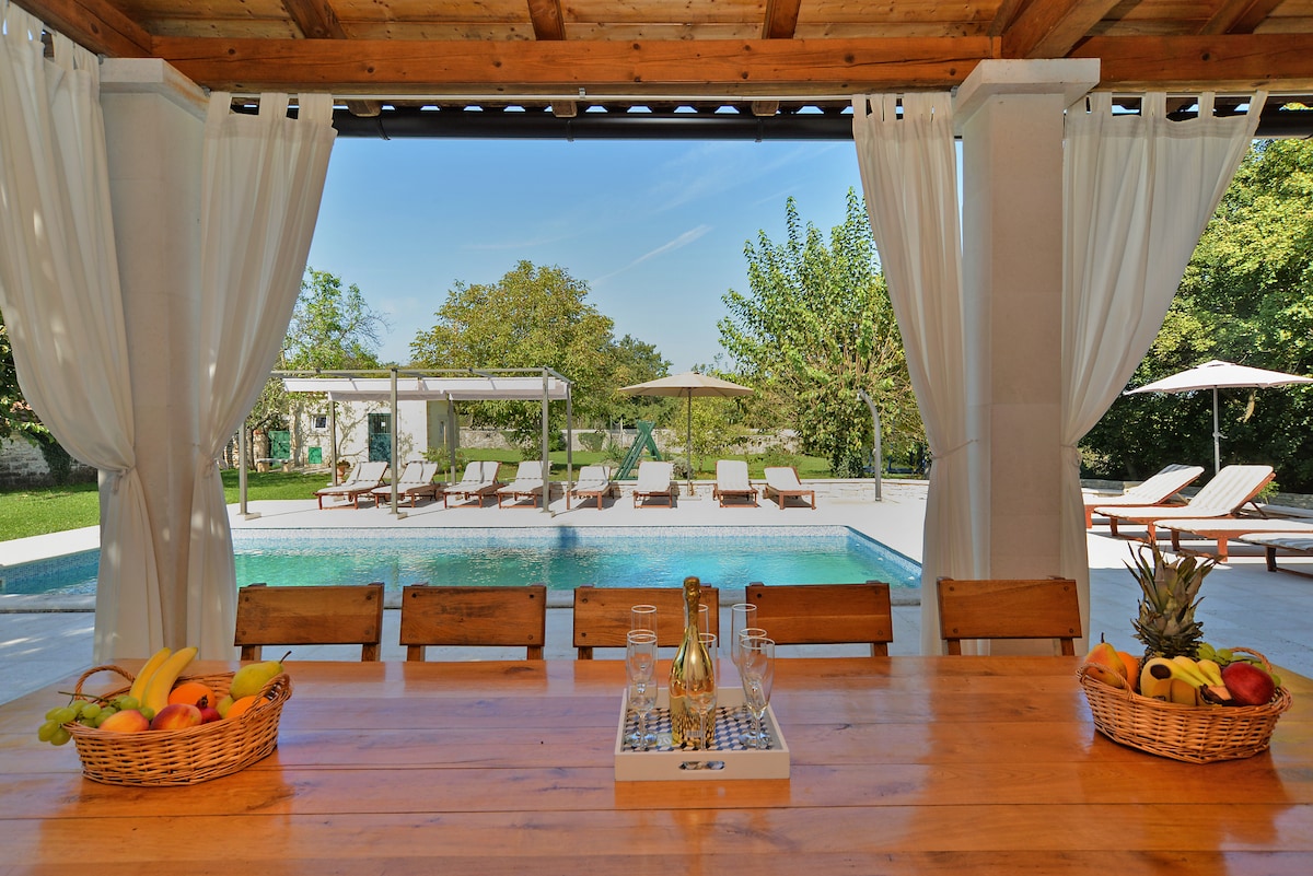 An outdoor dining area features a long wooden table, complemented by baskets of fresh fruit. Sheer curtains frame the view of a tranquil swimming pool surrounded by sun loungers and umbrellas, creating a serene atmosphere for relaxation.