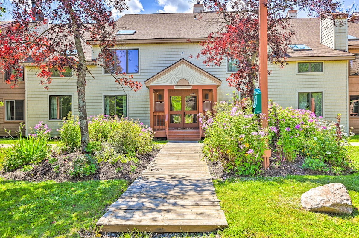 The entrance is framed by vibrant flowers and greenery, complemented by decorative trees displaying red leaves. A wooden pathway leads to the welcoming entrance, which features double doors and a covered porch, inviting guests into the building.