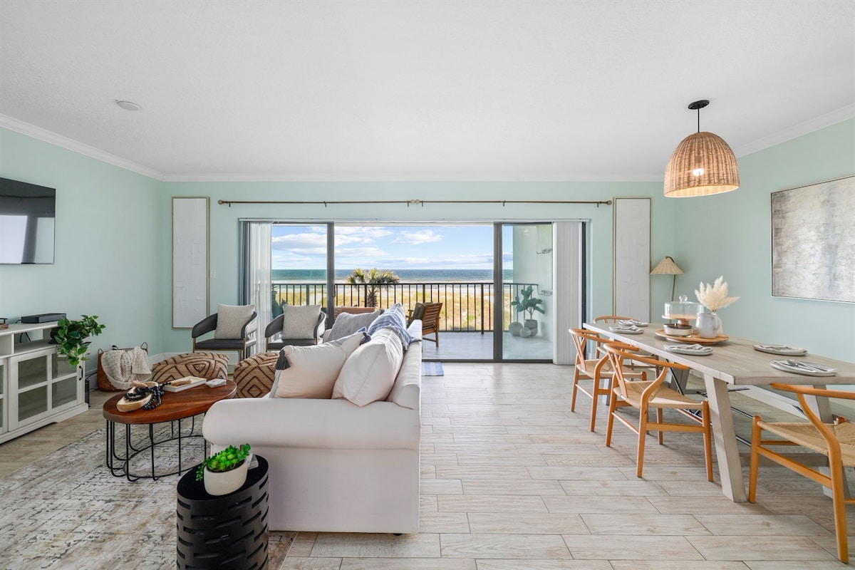 The open living area showcases a light and airy atmosphere with a comfortable sofa facing large sliding glass doors that provide views of the ocean. A dining table is set for meals, while decorative elements, like plants, add a touch of warmth to the coastal design.