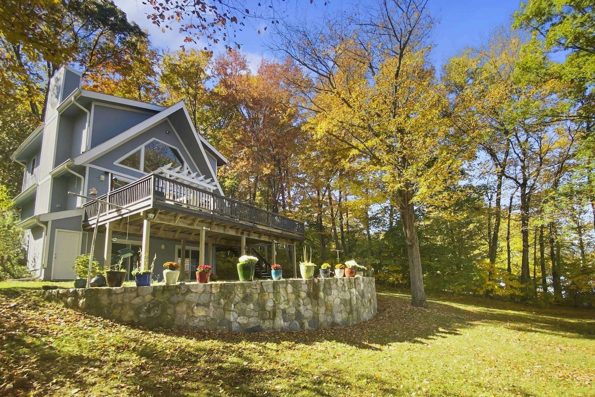 The exterior of a charming house is surrounded by vibrant autumn foliage. A spacious deck spans the front, with potted plants adding color against the stone wall. A grassy area in the foreground enhances the natural setting, inviting guests to explore the outdoors.
