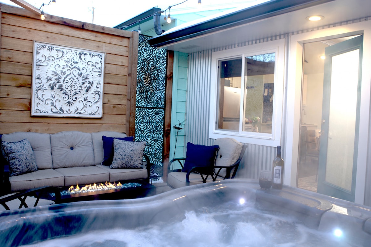 A private courtyard features a relaxing hot tub surrounded by seating and decorative elements. A cozy sofa with blue cushions offers comfortable lounging, while warm lighting from nearby café lights creates an inviting ambiance. The entrance to the studio is visible, enhancing accessibility.