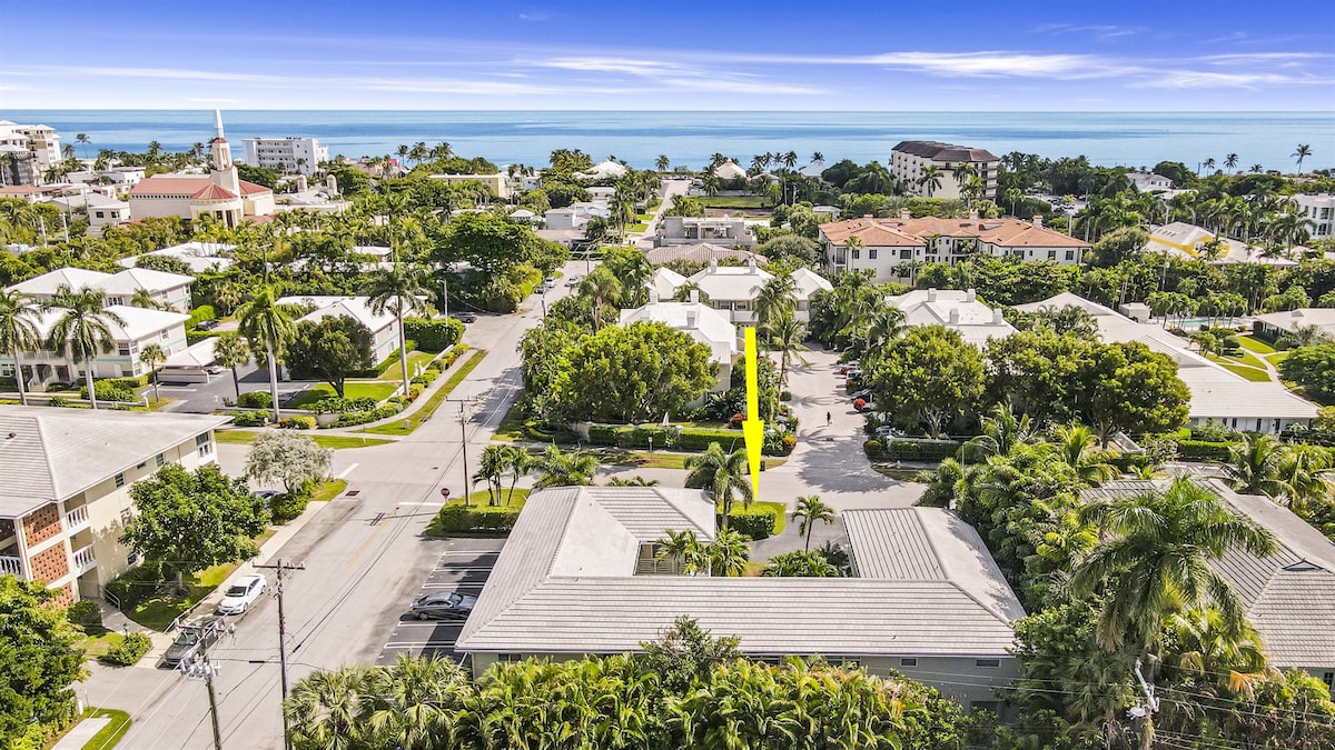 An aerial view showcases a vibrant neighborhood with lush greenery and palm trees. The coastline is visible in the distance, highlighting the proximity to the beach. The street layout is clearly seen, along with various residential buildings, creating a tranquil atmosphere.