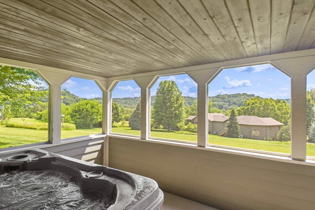 The screened-in gazebo features a private hot tub, surrounded by large octagonal windows that offer expansive views of the green landscape and nearby hills. The serene outdoor setting includes trees and well-maintained lawns, creating a peaceful atmosphere.
