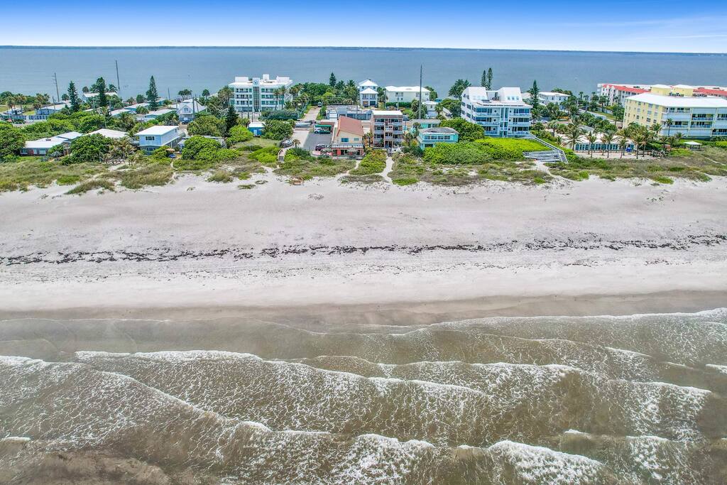 An aerial view of a beachfront property reveals white sandy shores meeting gentle waves. The surrounding area features various buildings and lush greenery, with the blue ocean extending into the horizon under a clear sky.