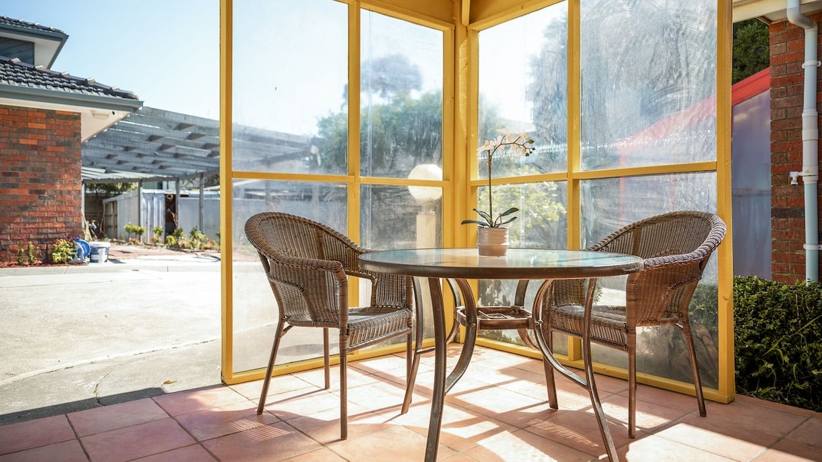 A covered outdoor area features a glass table surrounded by two wicker chairs. A potted orchid is displayed at the center of the table, while sunlight filters through the clear panels, providing a bright and open environment.