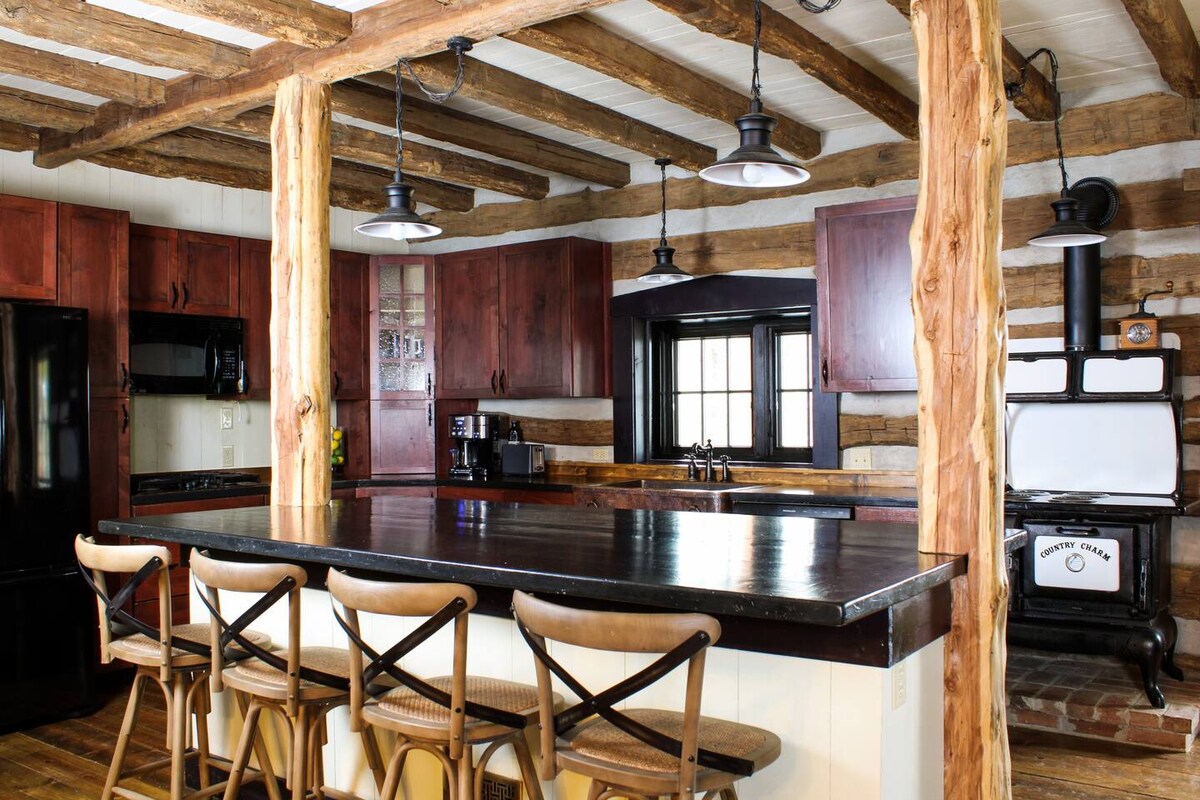 A rustic kitchen showcases modern amenities with wooden beams overhead and sleek cabinetry. A central island with seating for four is highlighted, with a black refrigerator and stove positioned nearby. Natural light is admitted through the window, illuminating the warm tones of the wood.