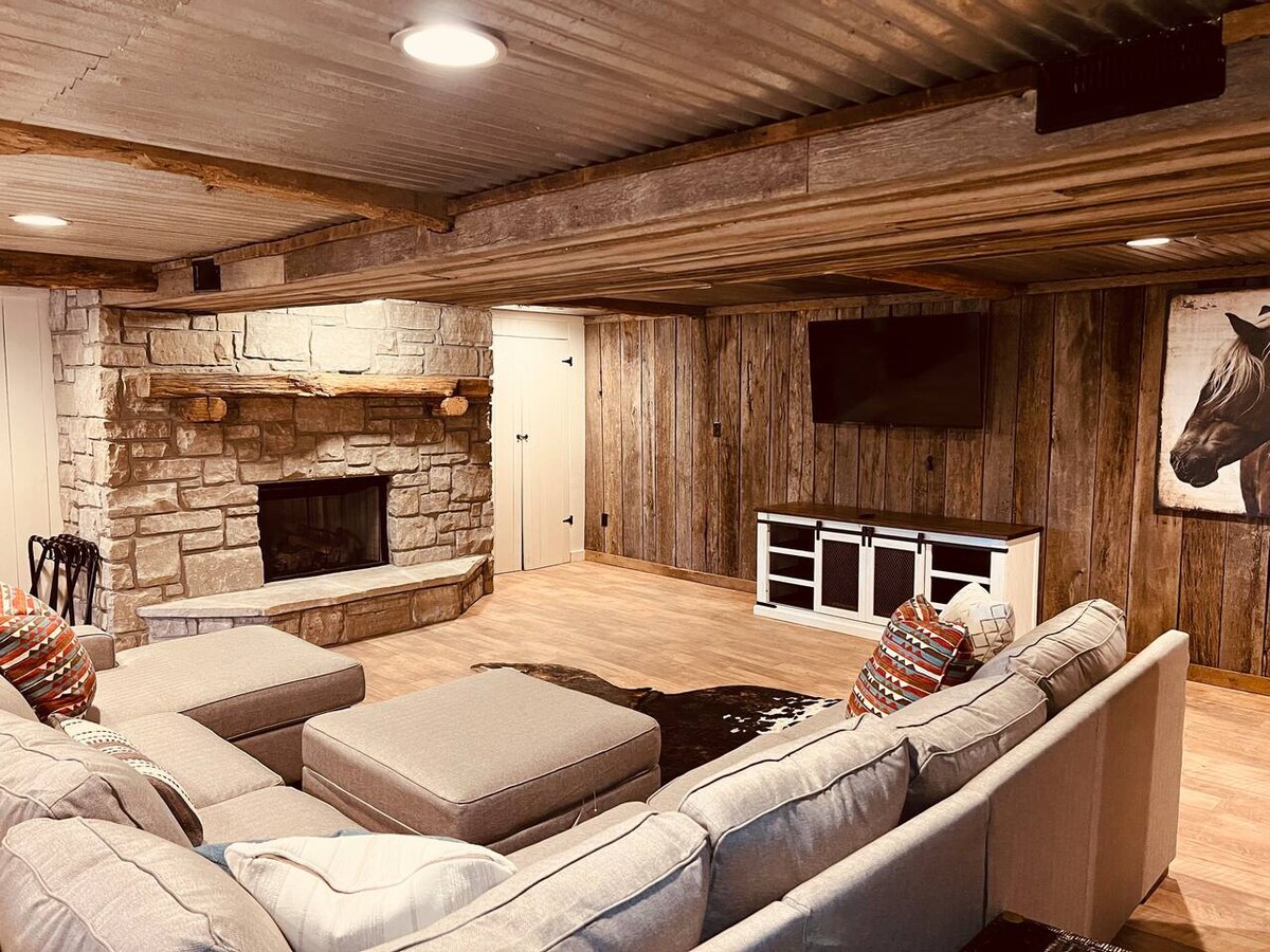 A spacious basement living area is furnished with a large sectional sofa and decorative cushions. A stone fireplace serves as a focal point against the wooden paneling. A wall-mounted TV is visible, and a piece of artwork featuring a horse adds character to the space.