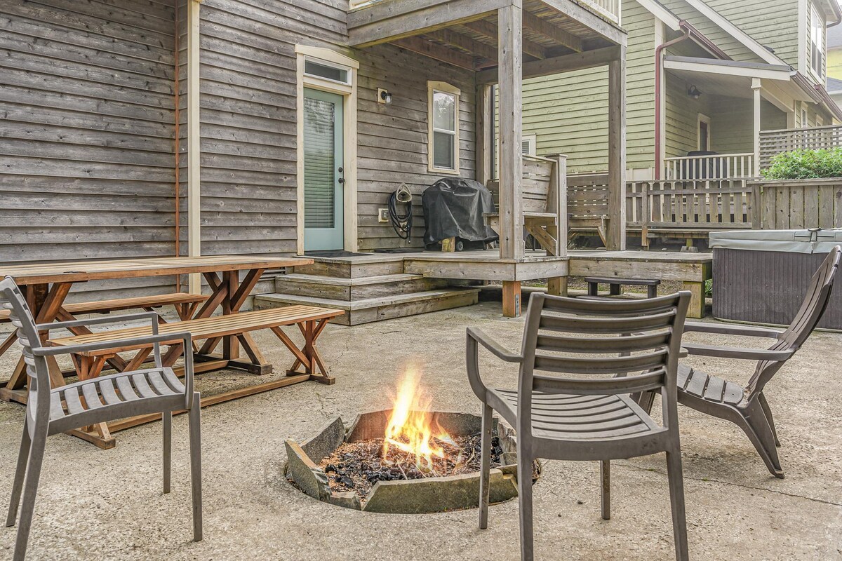 The outdoor space features a concrete patio with a built-in firepit surrounded by four chairs. A wooden dining table is positioned nearby, with an outdoor grill located under a covered area in the background, connecting to the home through a glass door.