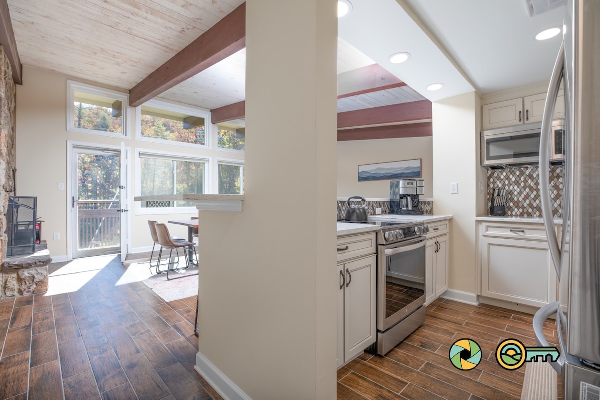 A modern kitchen is visible, featuring stainless steel appliances and tile backsplash. Natural light fills the space through large windows. A dining area with a table and chairs is positioned next to the kitchen, complemented by a stone fireplace adjacent to the seating area.