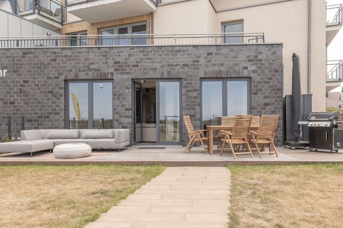 The outdoor area features a modern seating arrangement with a large sectional sofa and a wooden dining table surrounded by chairs. A grill is positioned nearby for outdoor cooking. Clear views of the lawn lead to the entrance of the stylish property.