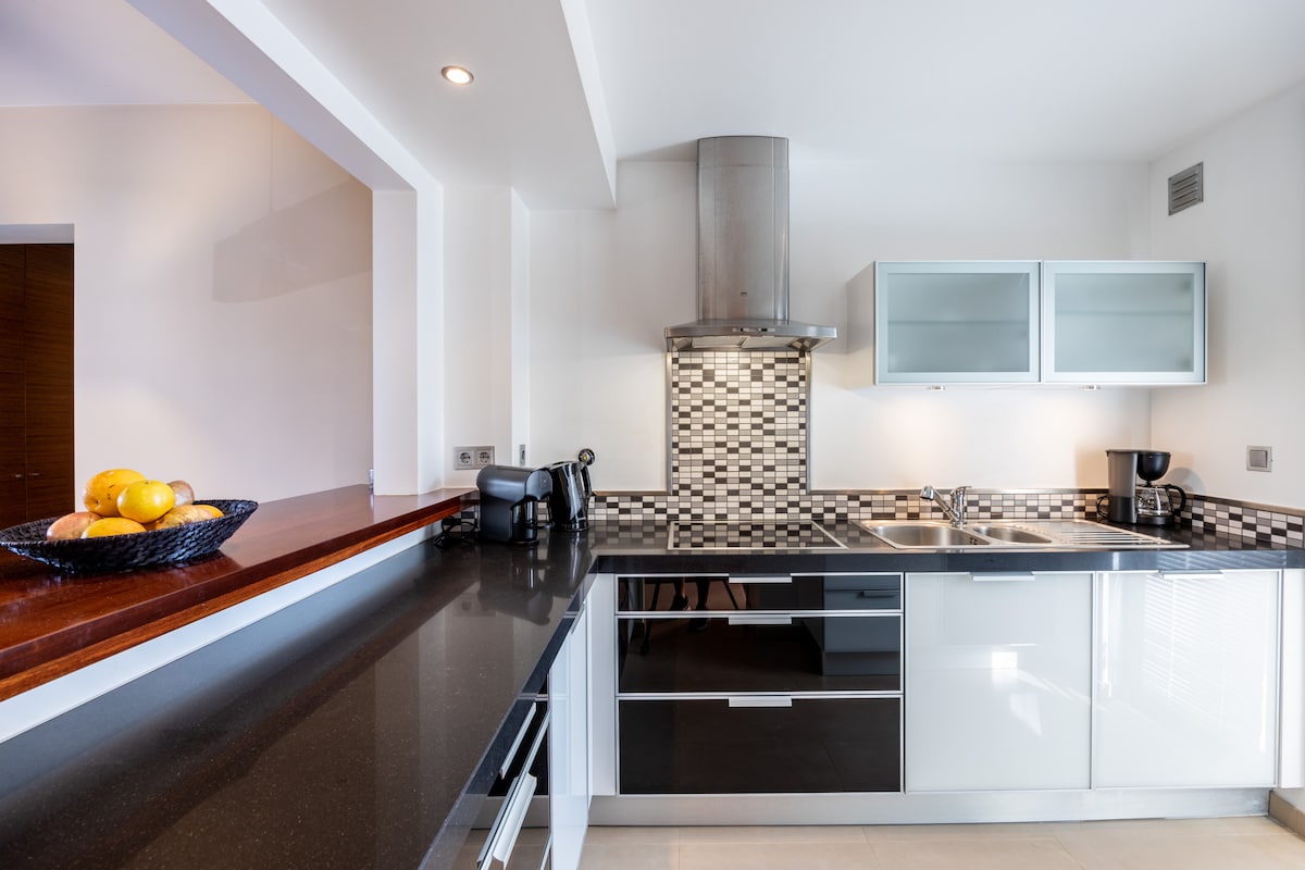 The kitchen presents a modern design with glossy cabinetry and a decorative backsplash in contrasting colors. A black countertop is equipped with appliances, including a kettle and coffee maker. The space features dual sinks, and natural light enhances the overall ambiance.