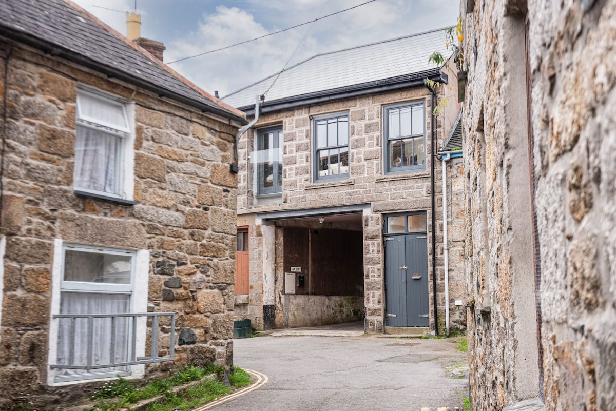 A quiet street scene features stone buildings with traditional architecture. The loft-style apartment has large windows and a garage door below, providing a welcoming entrance. Narrow pathways lead to the entrance, enhancing the sense of community in this charming Cornish neighborhood.