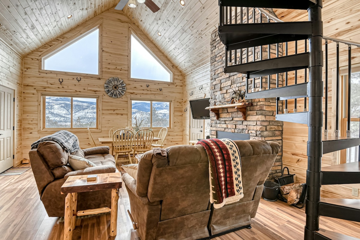 A relaxing living area features natural wood walls and large windows that frame stunning mountain views. Two comfortable sofas are arranged around a wooden coffee table, with a stone fireplace adding warmth. A spiral staircase leads to the loft area above.