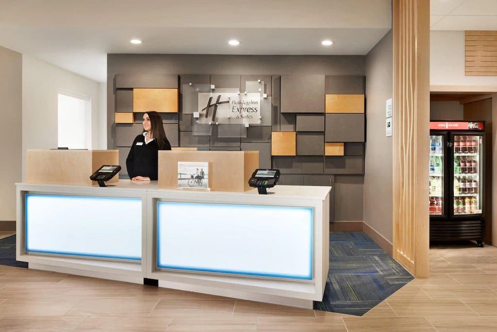 The reception area features a modern front desk with illuminated panels, complemented by a geometric wall design in neutral tones. A staff member is positioned behind the desk, and a vending machine is visible to the right, offering a variety of snacks and beverages.