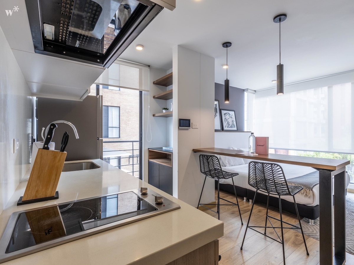 A modern kitchen is equipped with sleek appliances and countertop space. Stylish wire bar stools are positioned at a wooden dining table, bathed in natural light from large windows. Minimalist decor is evident throughout the space, enhancing the functional layout.