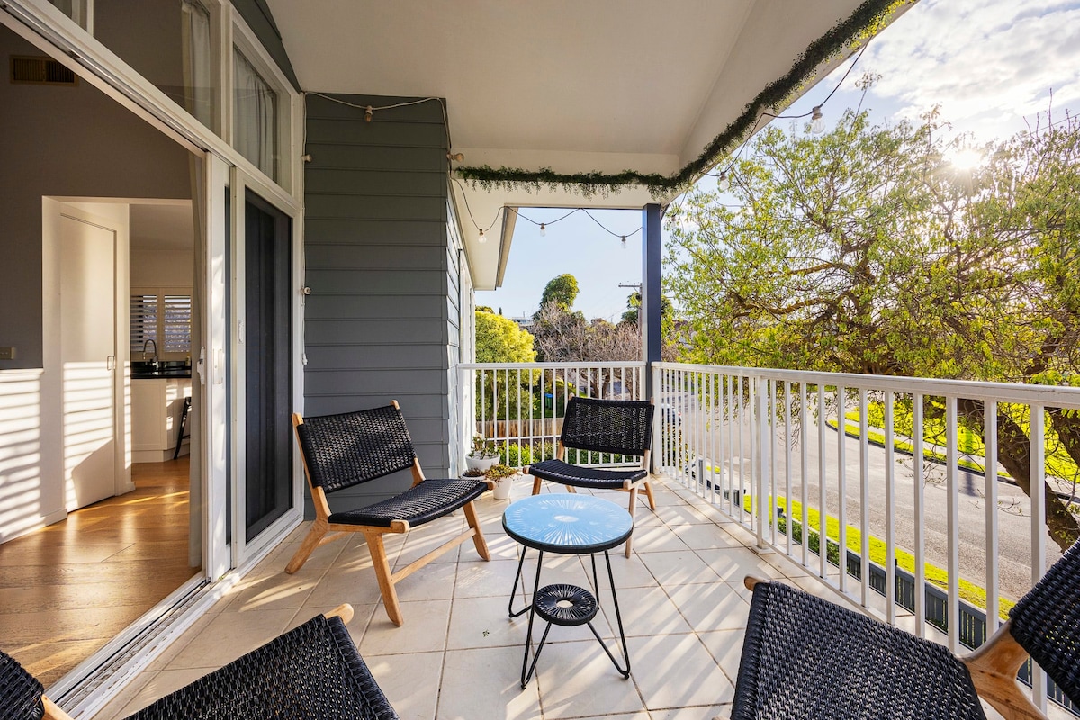 A covered balcony features several black wicker chairs arranged around a circular blue table. Large glass doors lead into the interior space. Soft sunlight illuminates the area, highlighting the green trees and distant street views beyond the railing.