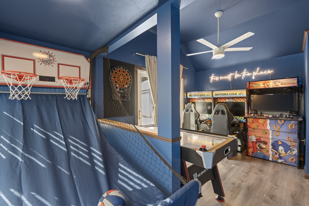 A dedicated game room features a Pop-A-Shot basketball game, a dartboard, and air hockey table. Two racing arcade cabinets stand against a vibrant blue wall, while a ceiling fan provides comfort. Natural light filters through a window adorned with light curtains.