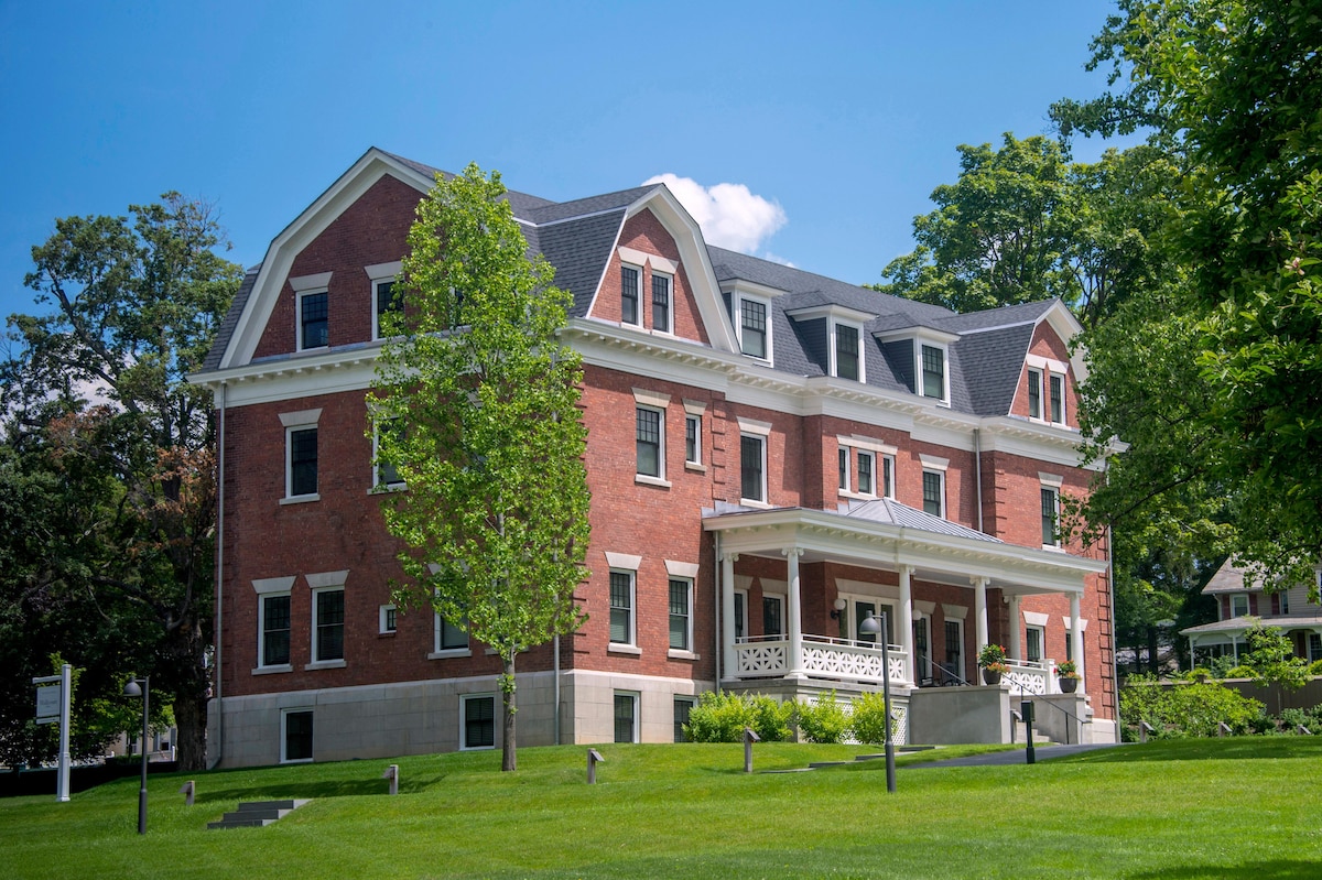 A red brick building with elegant architectural details is framed by green grass and trees. The expansive porch features white railings and is supported by tall columns, creating an inviting entrance. Bright blue skies and scattered clouds add to the serene atmosphere.