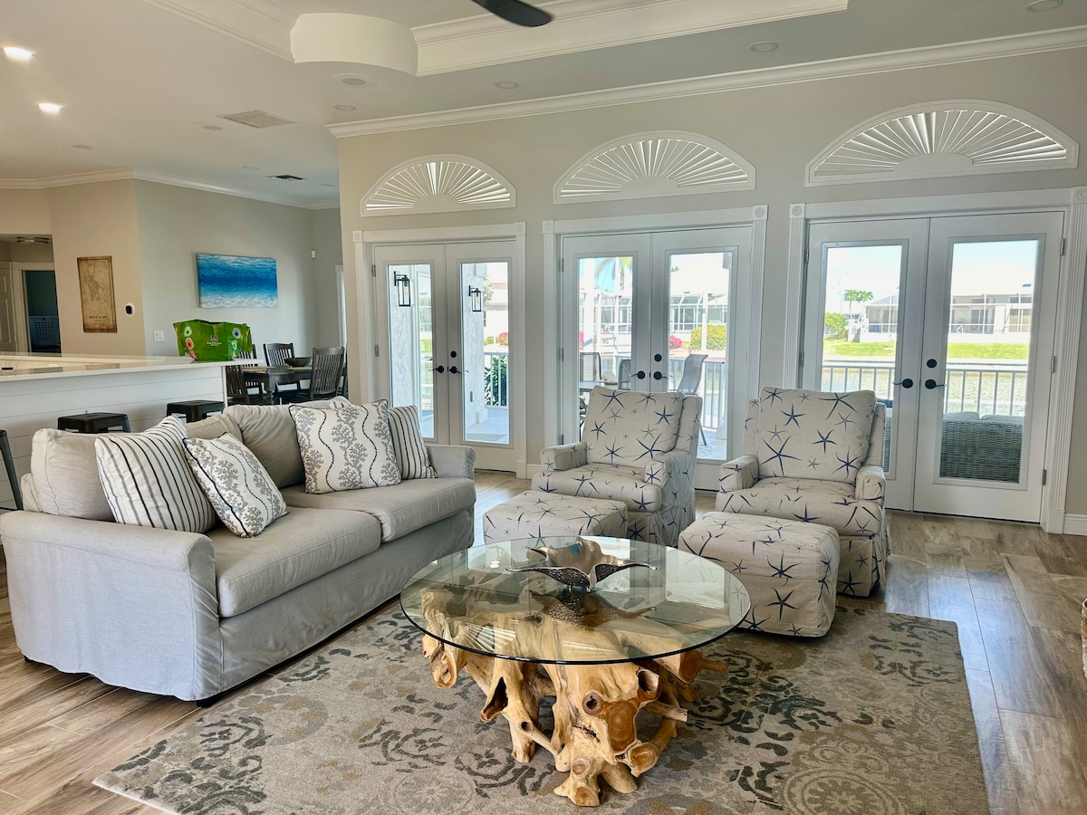 A comfortable living area features a light grey sofa with decorative pillows, flanked by two upholstered armchairs. A round glass coffee table rests on a patterned area rug, with large glass doors providing views of the waterfront and allowing natural light to fill the space.