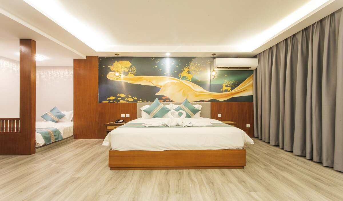 The room features a spacious bed adorned with decorative pillows and a unique wall mural depicting serene scenes. Soft lighting enhances the ambiance, while light-colored wooden flooring complements the overall design. Dark curtains frame the window, offering options for natural light control.