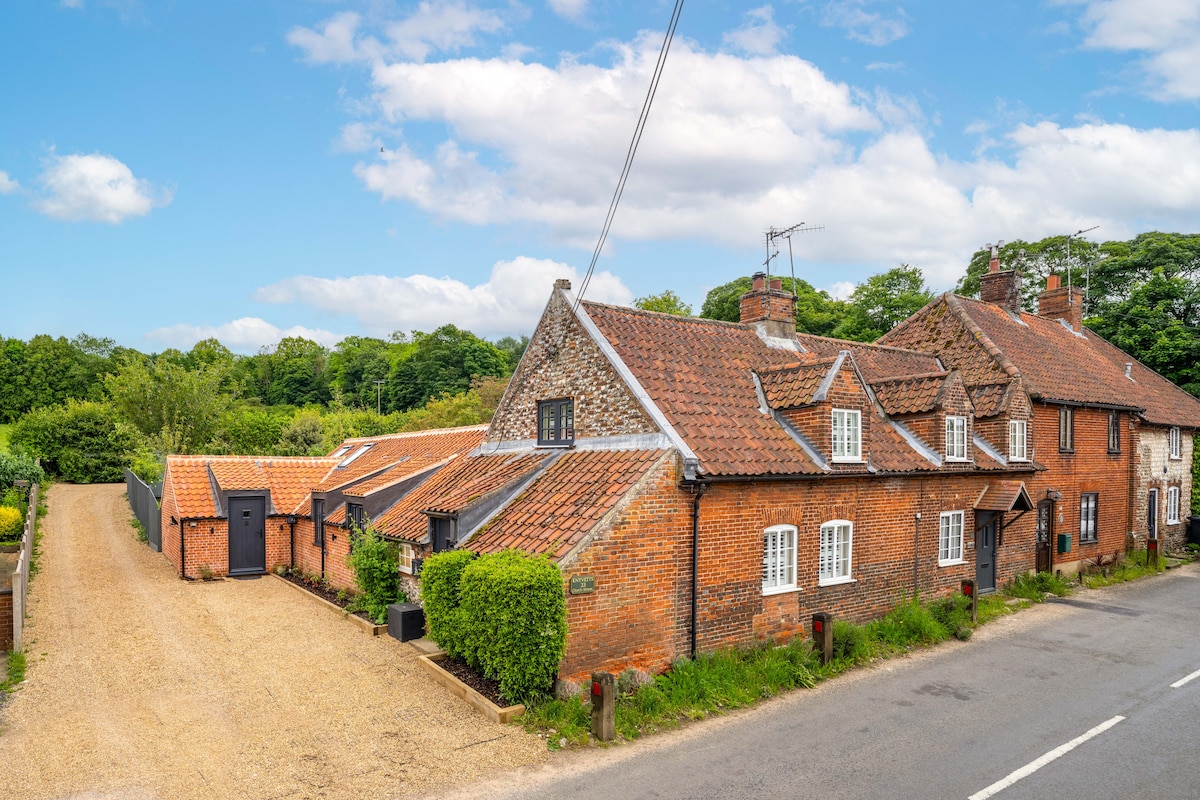 A charming three-bedroom cottage is showcased, featuring a mix of red brick and flint with a traditional tiled roof. The gravel driveway leads to the entrance, surrounded by neatly trimmed hedges and green trees in the background, providing a sense of seclusion and comfort.
